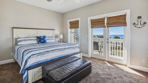 Un dormitorio con cama, un banco, ventilador de techo y vistas al océano a través de amplios ventanales.