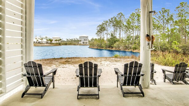 A covered outdoor seating area with Adirondack chairs facing a lake, surrounded by trees and buildings. - 3BR Home @ Summer Salt • Sleeps 9 • Resort Pool & Near Beach (Orange Beach)