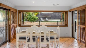 A dining area with a table set for four, a view of the garden through a window, and wooden shutters on the sides.