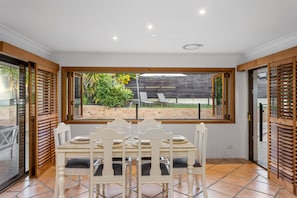 A dining area with a table set for four, a view of the garden through a window, and wooden shutters on the sides. - 4BR Family Oasis w/ Pool Modern and Spacious (Robina)