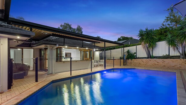 A swimming pool with a covered patio area, a brick wall, and a white fence.