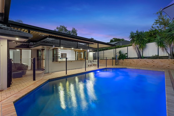 A swimming pool with a covered patio area, a brick wall, and a white fence. - 4BR Family Oasis w/ Pool Modern and Spacious (Robina)