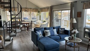 A spacious living room with a blue sectional sofa, a dining area with a wooden table and chairs, a spiral staircase, and large windows offering views of the outdoors. - Private Dock on Pine Lake – Sleeps 15 – Family Lakefront Retreat (Plainwell)