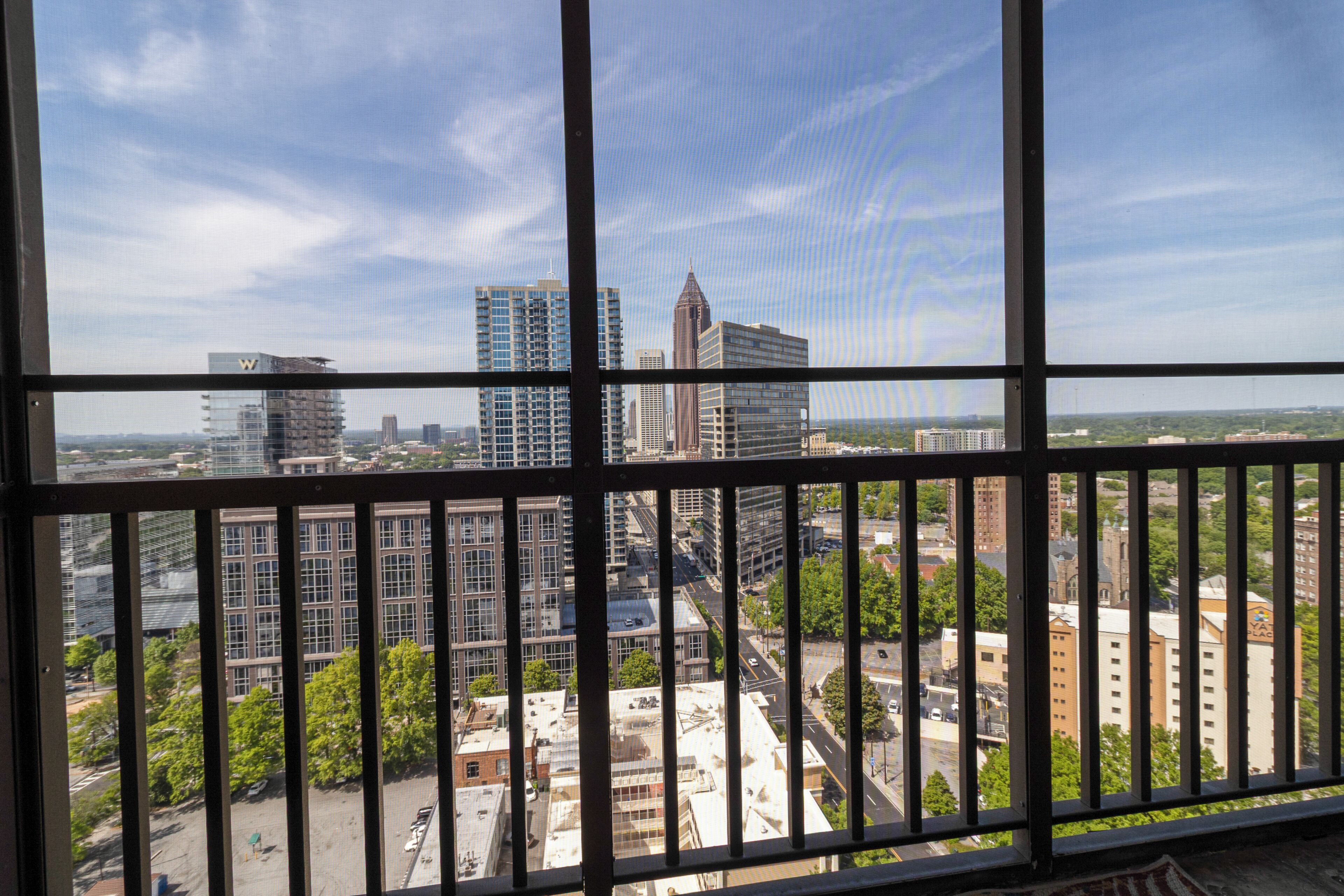 A cityscape through a window with a metal railing, featuring high-rise buildings, a prominent tower, and a mix of greenery and urban structures.