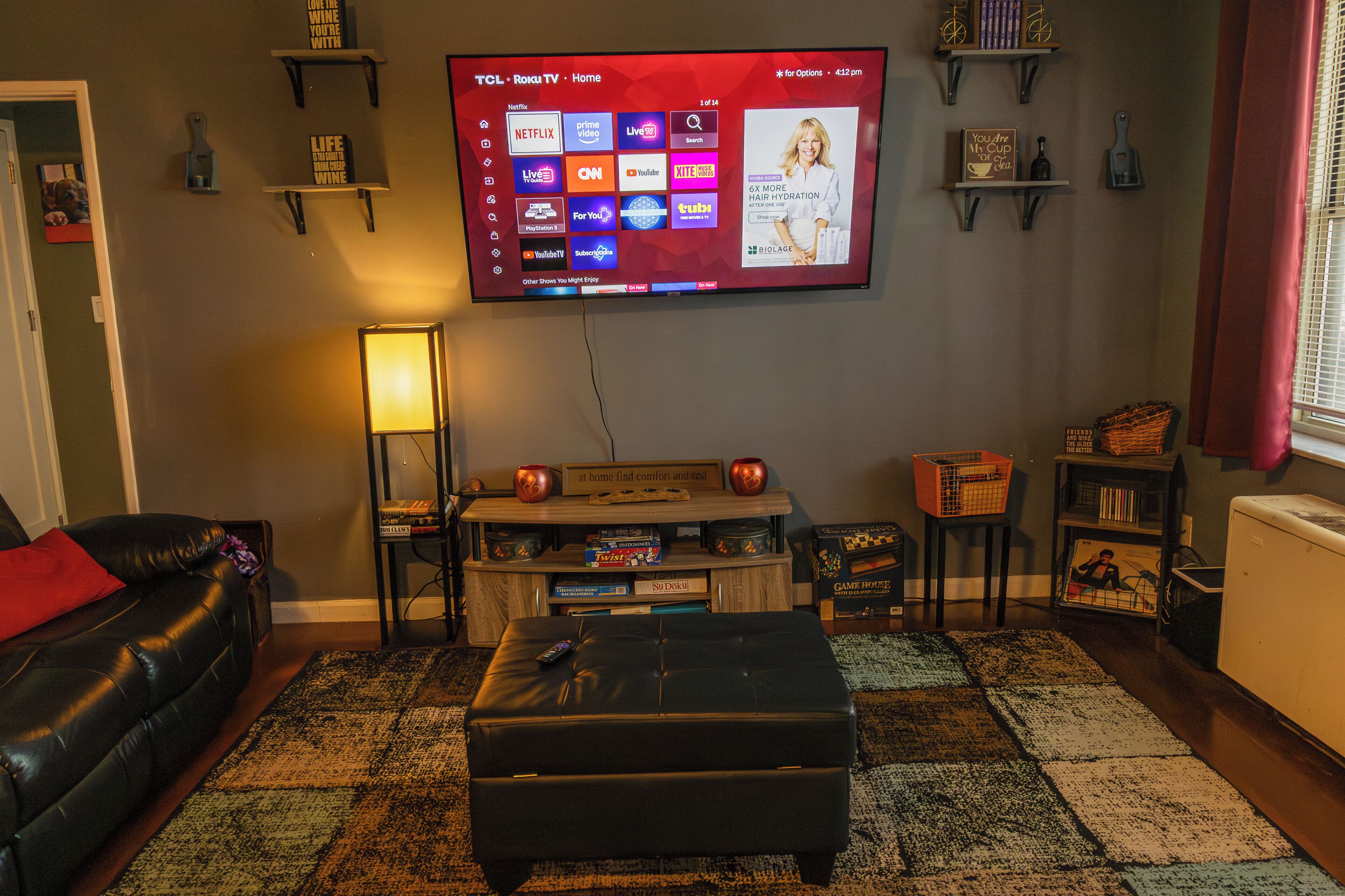A living room with a flat-screen TV mounted on the wall, a black leather sofa, a black ottoman, and a patterned rug.