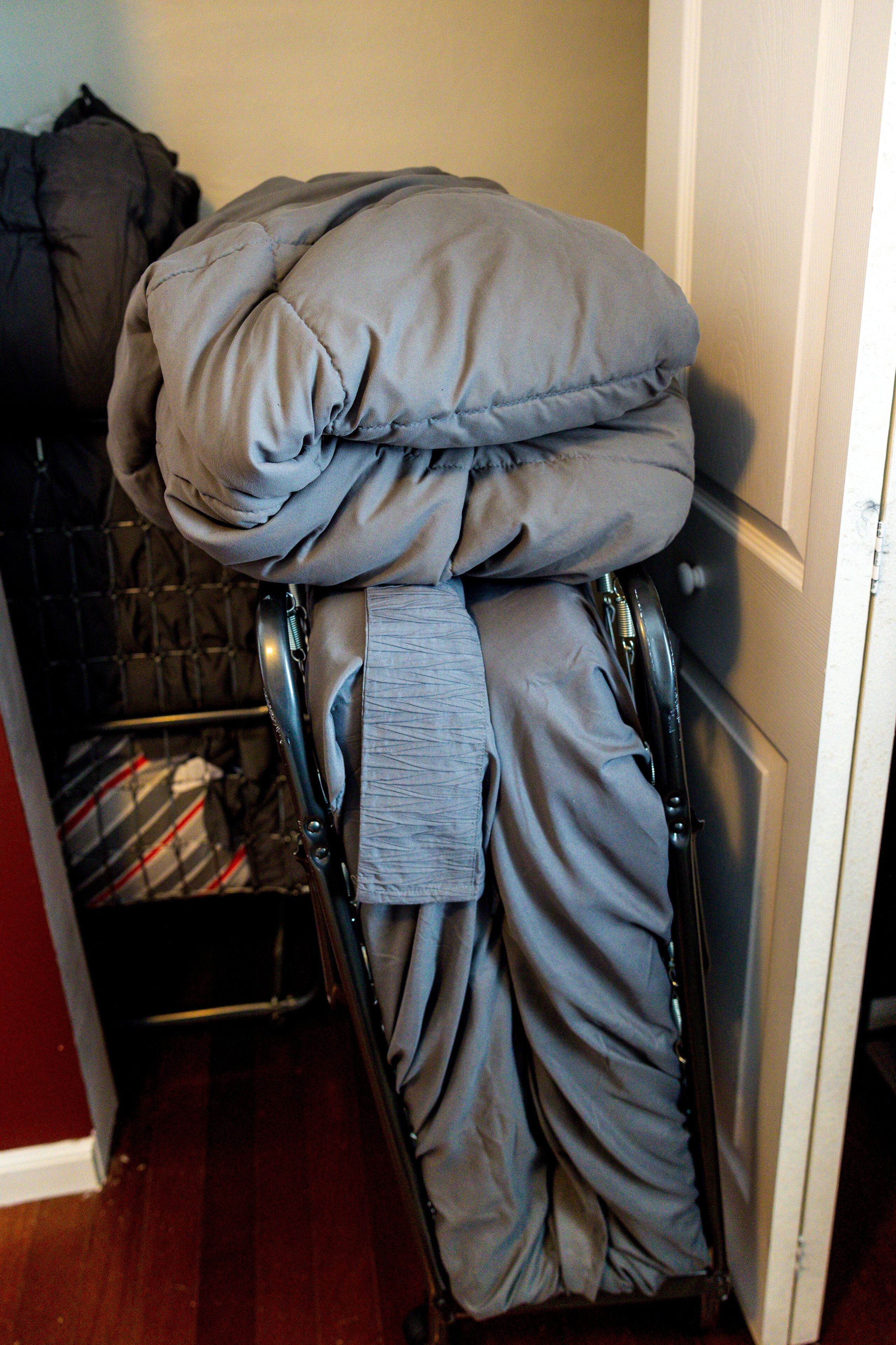 A compact bed with a grey blanket and pillow, a metal frame, and a striped blanket in the background.
