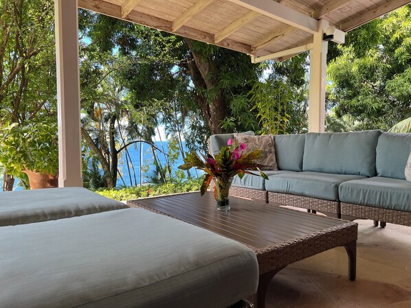 Un espacio de estar techado al aire libre con un sofá y una mesa, rodeado de abundante vegetación y vistas al mar.