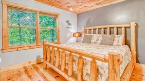 A wooden cabin bedroom with a bed, a window with a view of trees, and a deer wall decal. - Relaxing Helen, Ga Cabin Min from Helen, Sleeps 12 with Hot Tub & Firepit! 
     (Sautee Nacoochee)