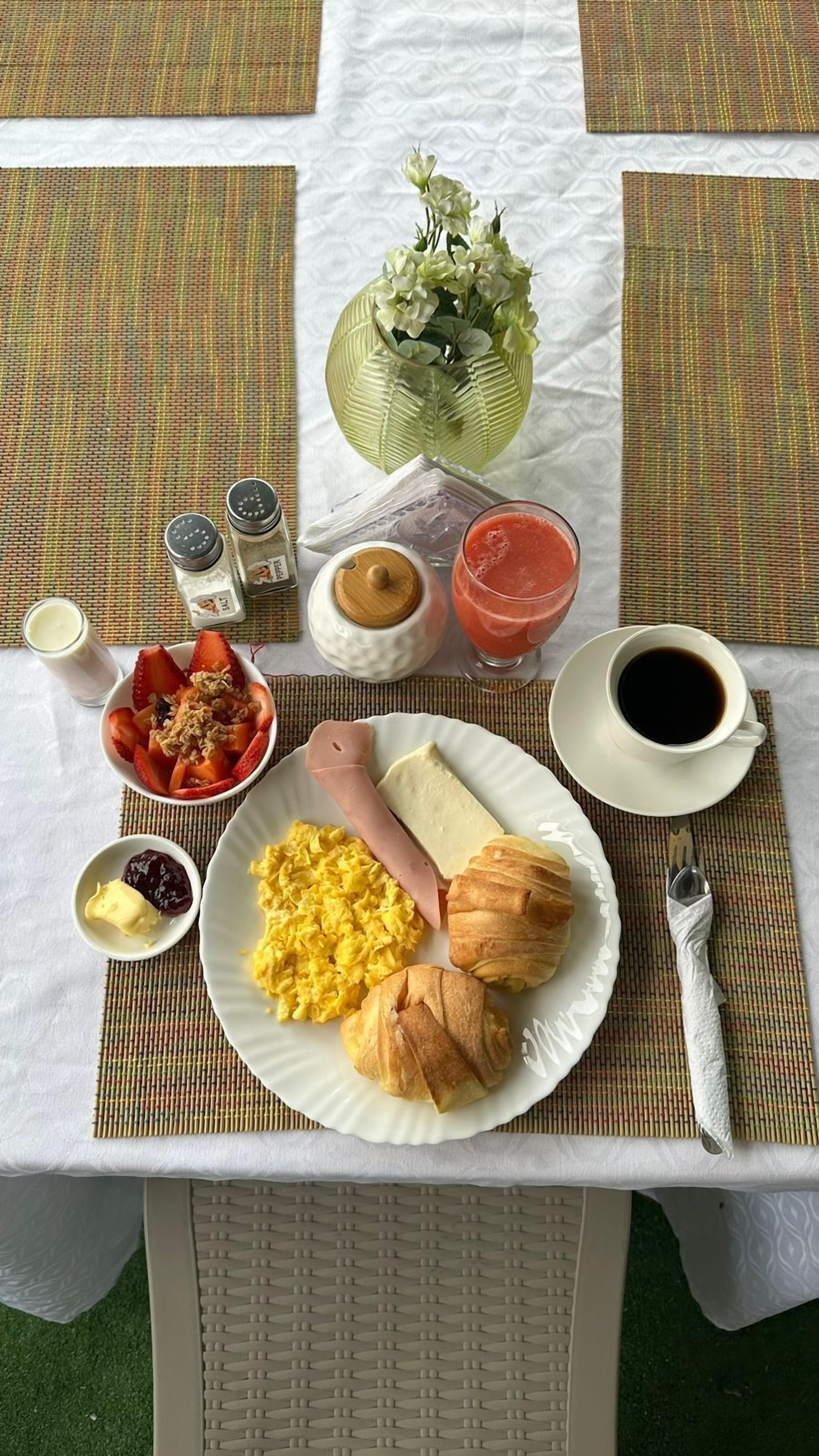 A breakfast table with a plate of scrambled eggs, ham, croissants, and a bowl of fruit. There are also condiments, a glass of juice, and a cup of coffee.
