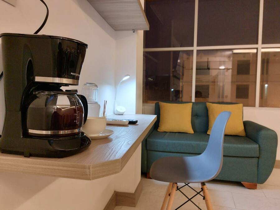 A modern living space with a coffee maker on a wooden table, a teal sofa with yellow cushions, and a window with a view of the city at night.