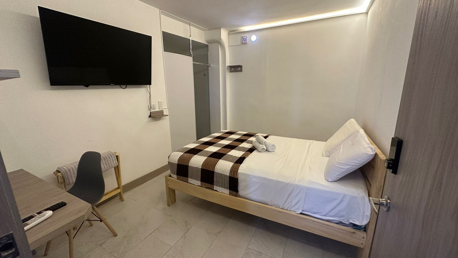 Una cama individual con una manta a cuadros, una mesita de noche con un control remoto, una silla y un televisor montado en la pared.