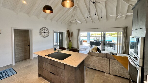 A spacious kitchen with a large island, modern pendant lights, and a view of an outdoor pool area. - Spacious Waterfront Sanctuary (Jolly Harbour)