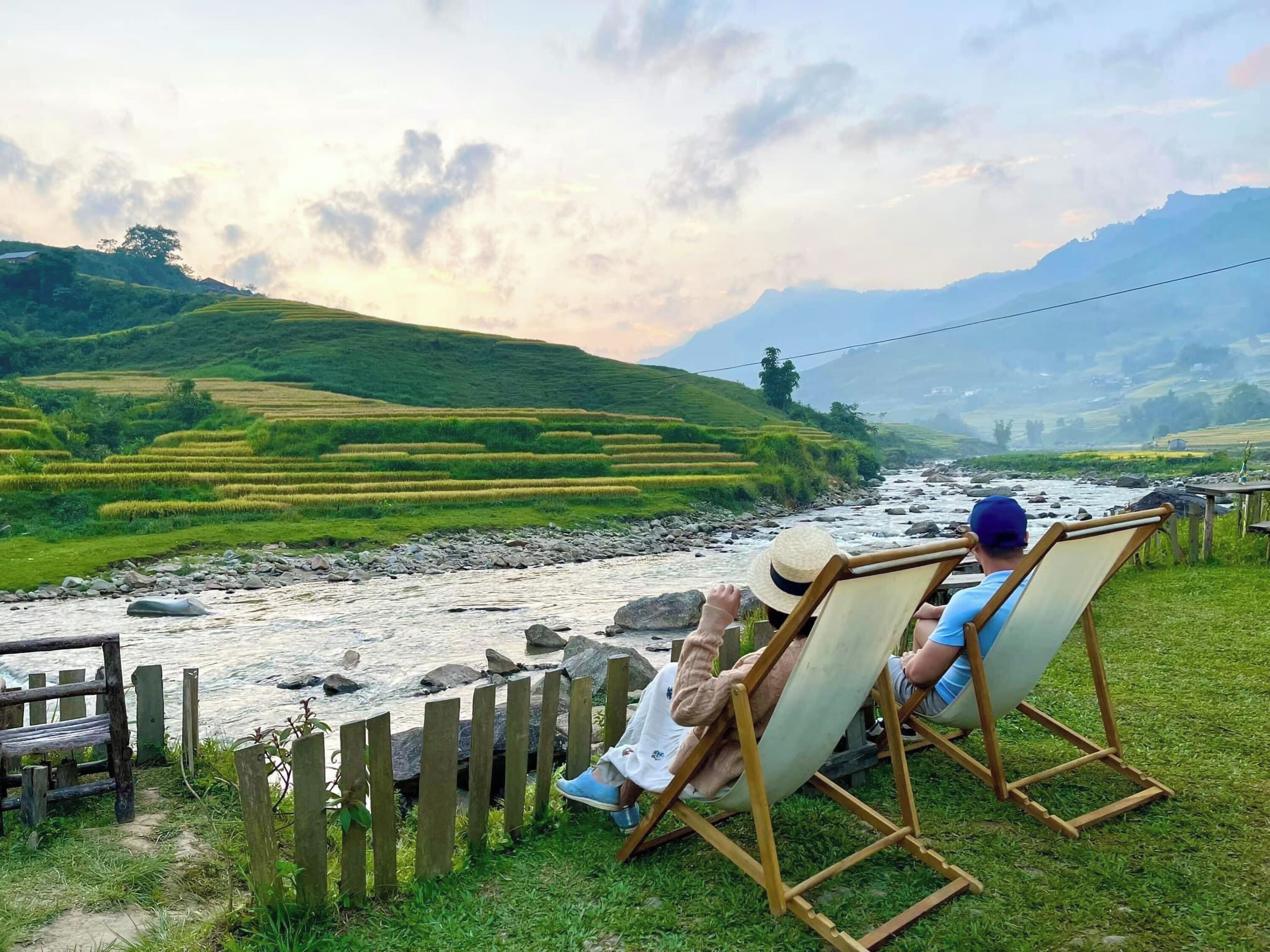 Two individuals relaxing on wooden deck chairs by a river with terraced fields in the background.