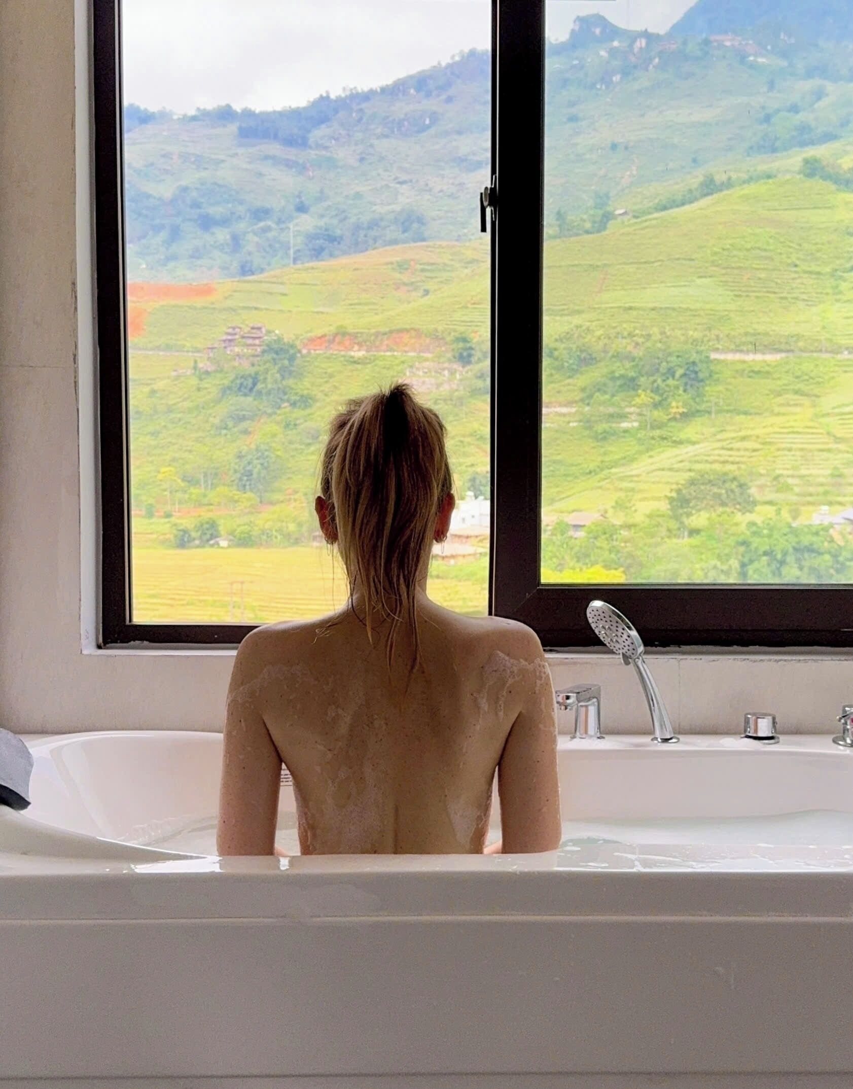 Una persona sentada en una bañera, con vistas a un paisaje verde a través de una ventana.