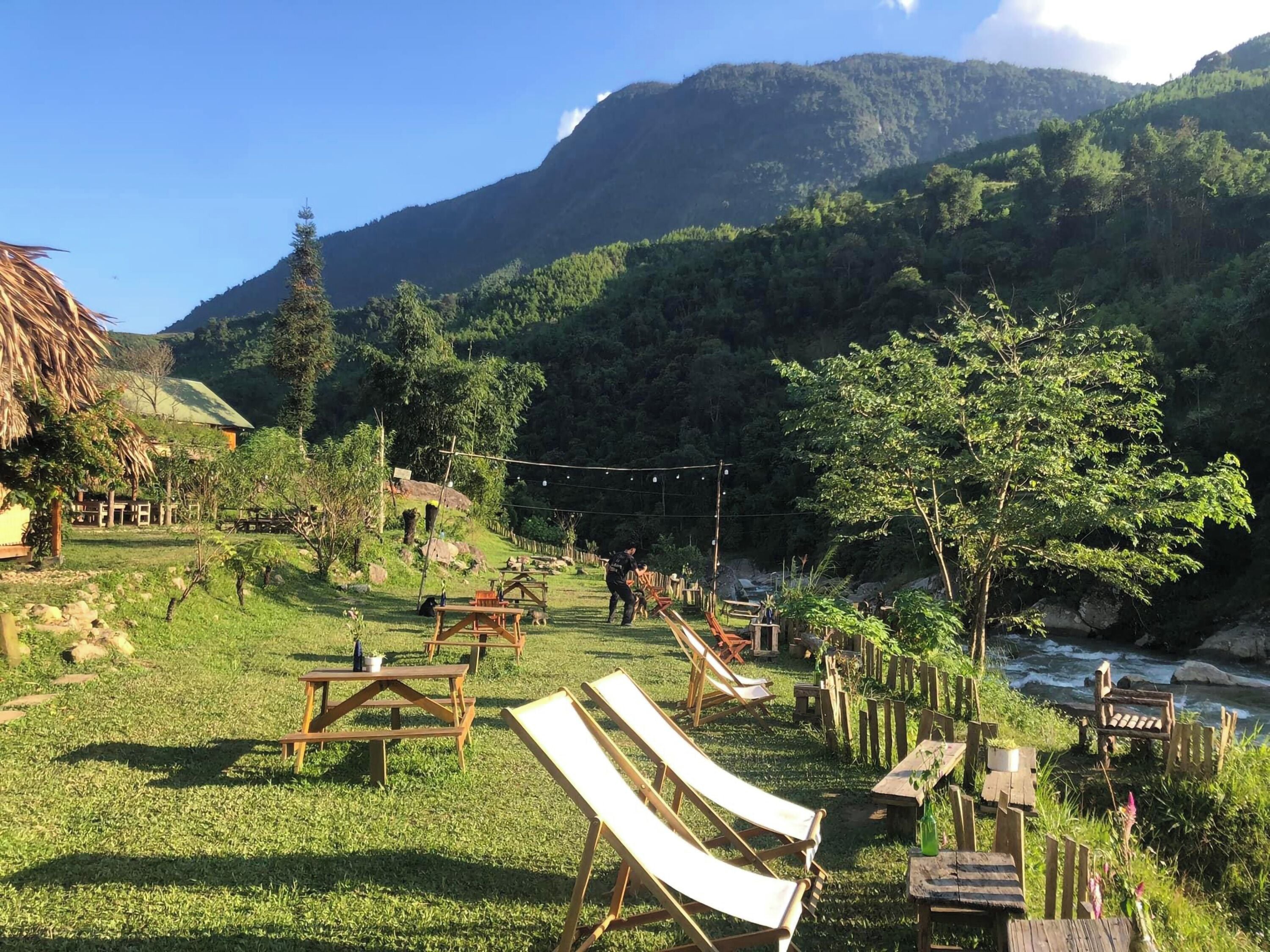 Un entorno rústico al aire libre con mesas de picnic y sillones de descanso, rodeado de exuberante vegetación y un río.