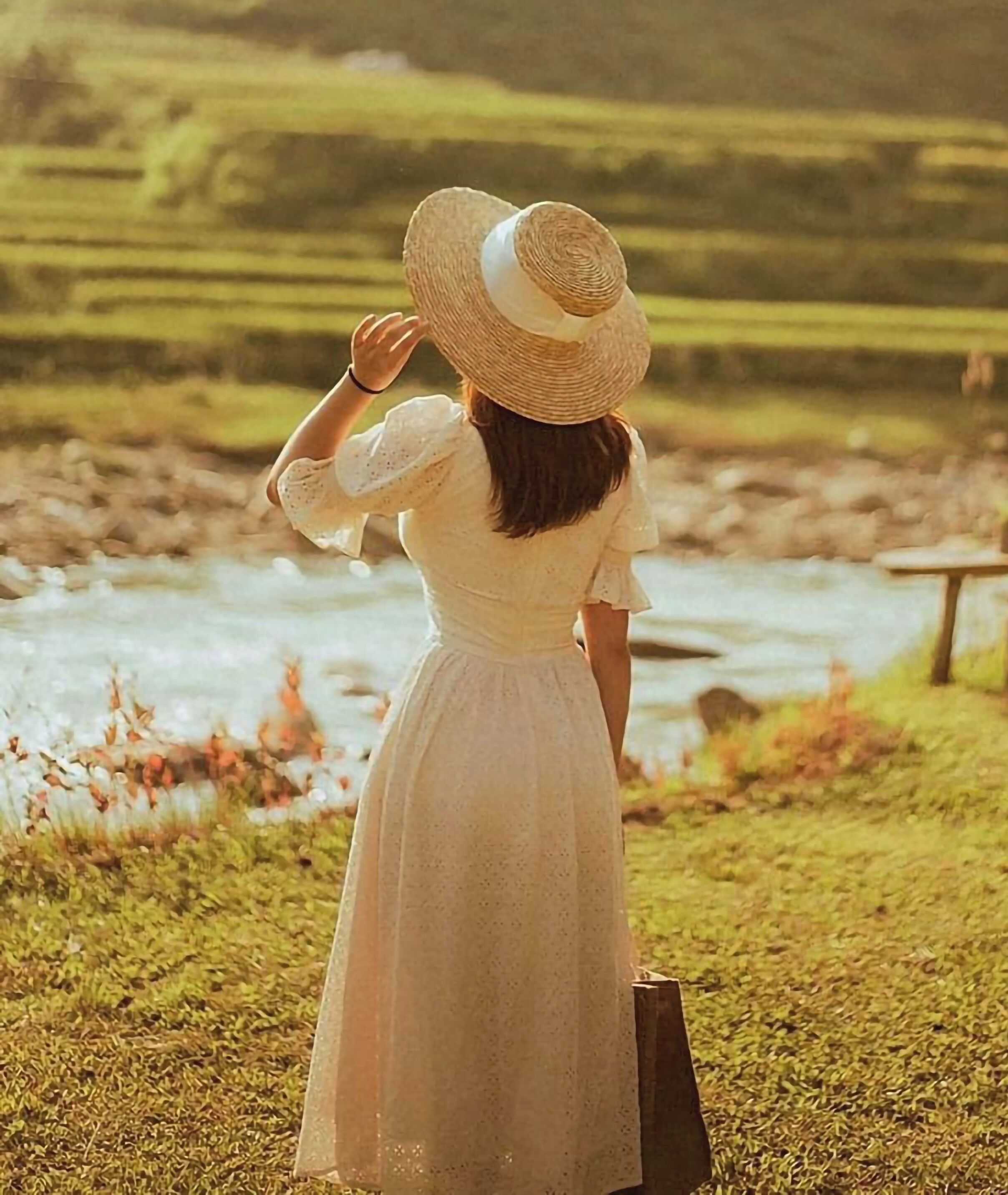 A person in a white dress standing by a river with a grassy area and a wooden structure in the background.