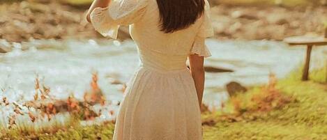 A person in a white dress standing by a river with a grassy area and a wooden structure in the background.
