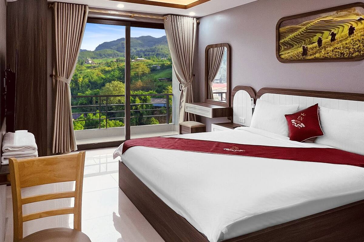 A hotel room with a large bed, a wooden chair, a desk, and a view of a green landscape through a sliding door.