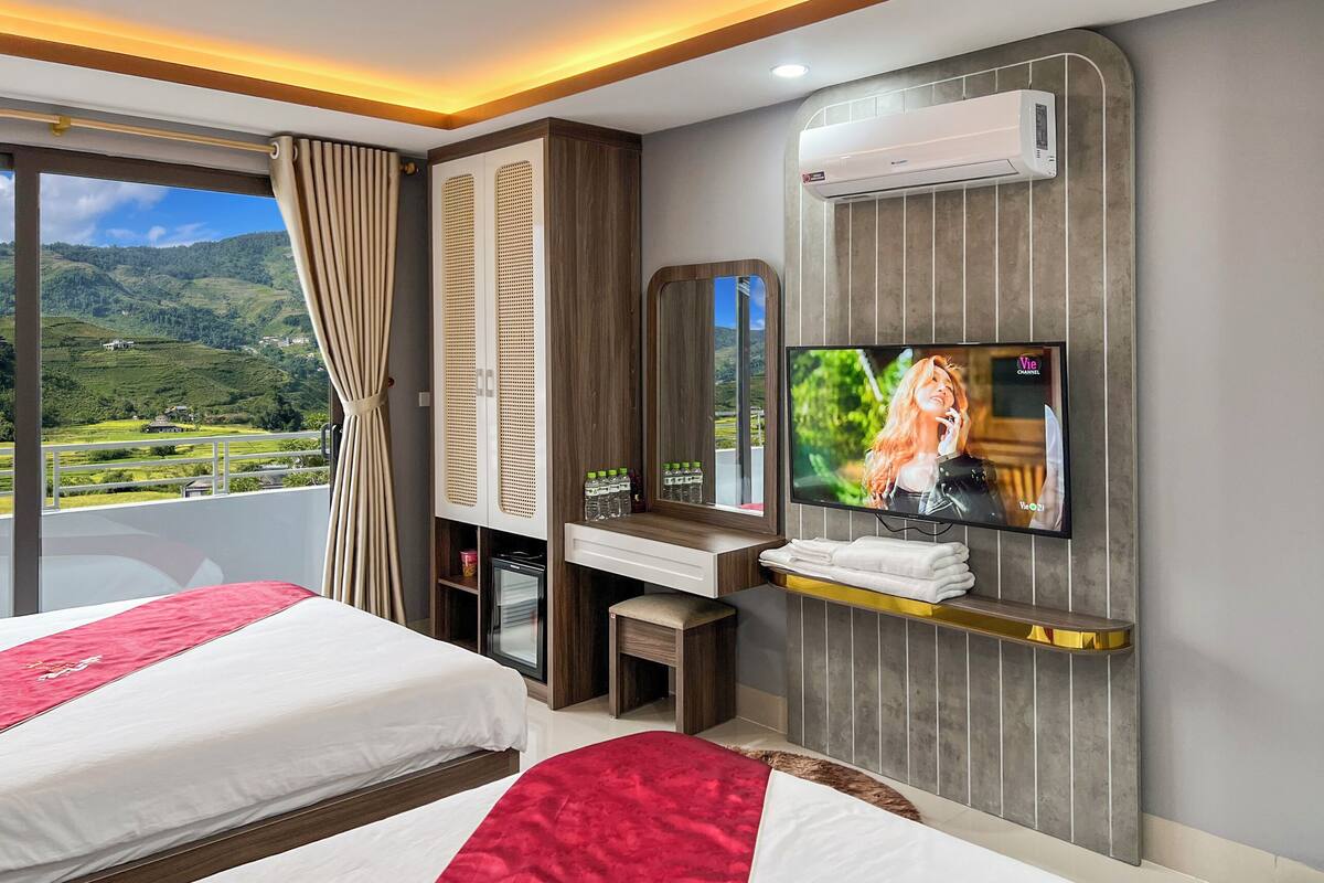 A hotel room with two beds, a TV, a minibar, and a view of a green landscape.