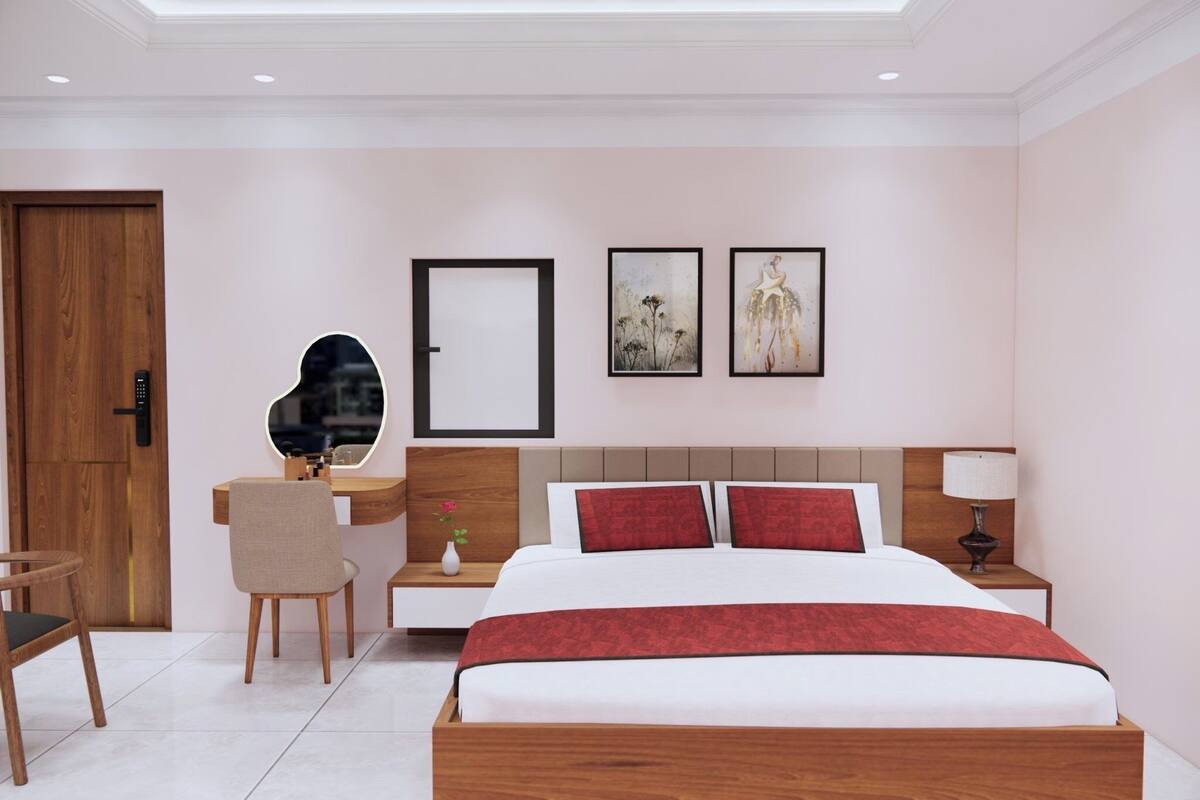 A modern hotel room with a wooden bed, a desk with a chair, a bedside table with a lamp, and two framed artworks on the wall.