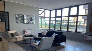 Un salon moderne avec un canapé, un fauteuil et une table basse, de grandes fenêtres donnant sur l’extérieur et des œuvres d’art abstraites accrochées au mur.