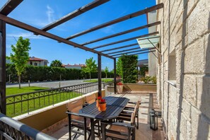 A covered outdoor patio with a table and chairs, a view of a road and houses, and a stone wall.