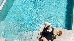 Une femme, vêtue d’un maillot de bain noir et d’un chapeau, est assise au bord d’une piscine, un bol de fruits à ses côtés.