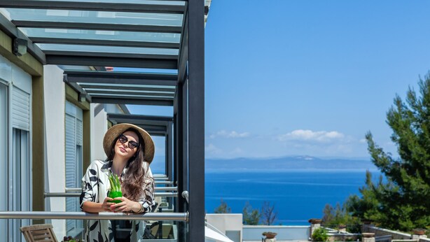 Un balcon donnant sur la mer, une personne tenant un verre, et la façade d’un immeuble moderne.