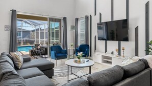 Ein modernes Wohnzimmer mit Blick auf einen Pool, graue Sofas, blaue Sessel, ein Flachbildfernseher und ein kleiner Tisch mit einer Vase.