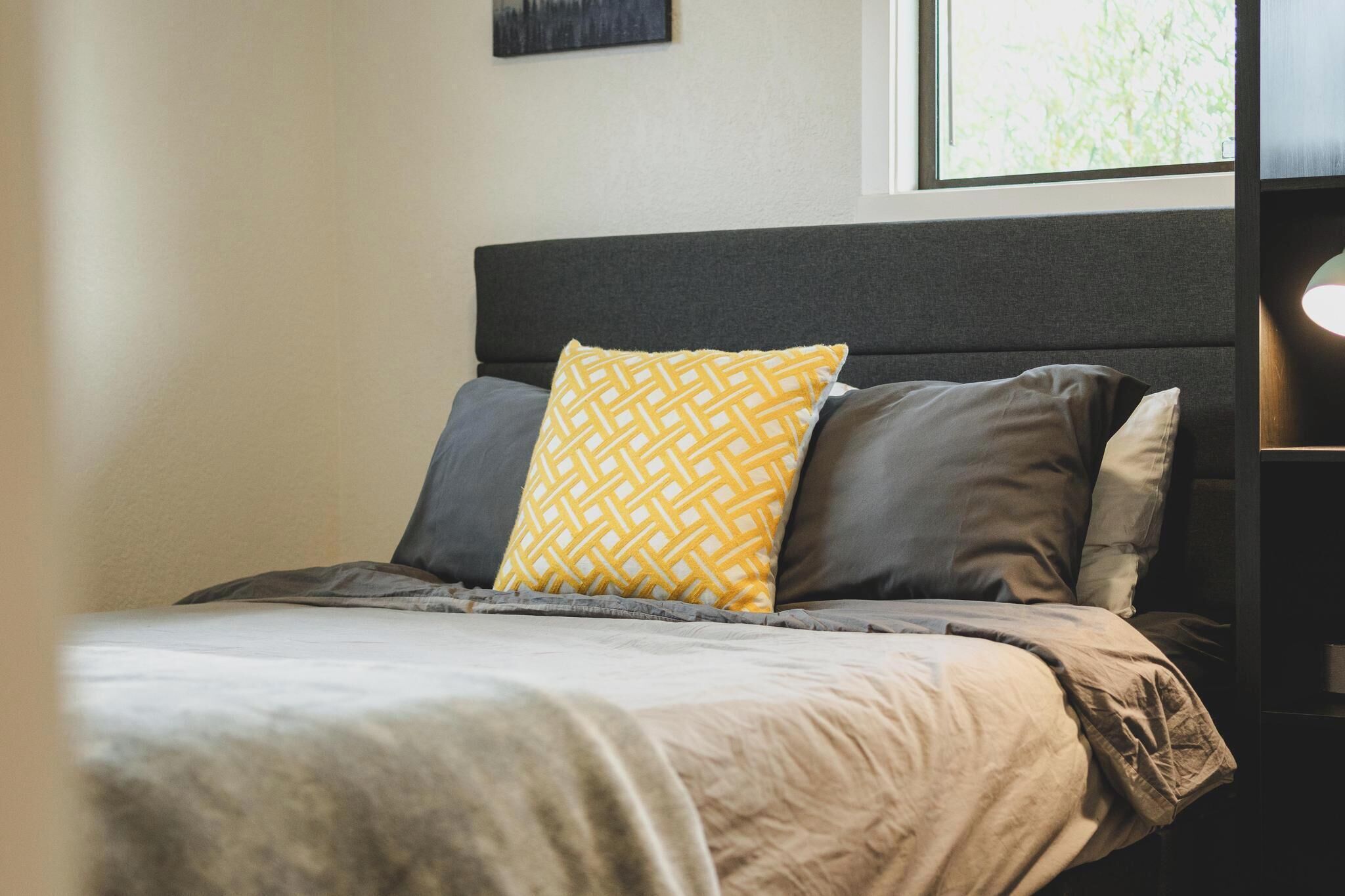Una cama bien hecha con un cabecero gris, un cojín amarillo estampado y un cojín gris.