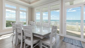 Un comedor con mesa de vidrio y sillas blancas, ventanas amplias con vista a la playa, y suelo de madera.