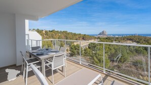 Un balcón con mesa dispuesta para comer, con vistas a un paisaje costero con árboles y una ciudad distante, además de una formación rocosa.
