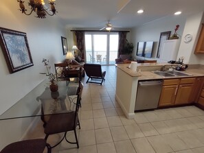 A modern kitchen with a dishwasher, a dining area with a glass table, and a living room with a sofa and a TV.