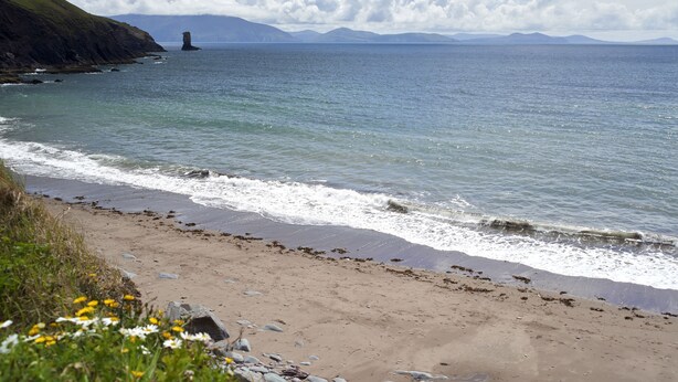 Une plage bordée d’une côte rocheuse, avec des vagues et un phare visible au loin.