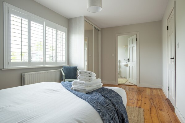 A bedroom with a large bed, a window with blinds, a door leading to a bathroom, and a stack of white towels on the bed.