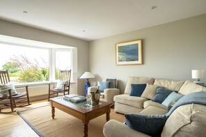 A living room with a beige sofa, a wooden coffee table, and a rocking chair. There is a window with a view of greenery outside.