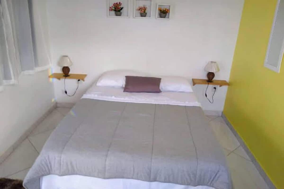 A bedroom with a bed, two bedside tables with lamps, a window with curtains, and three framed pictures on the wall.