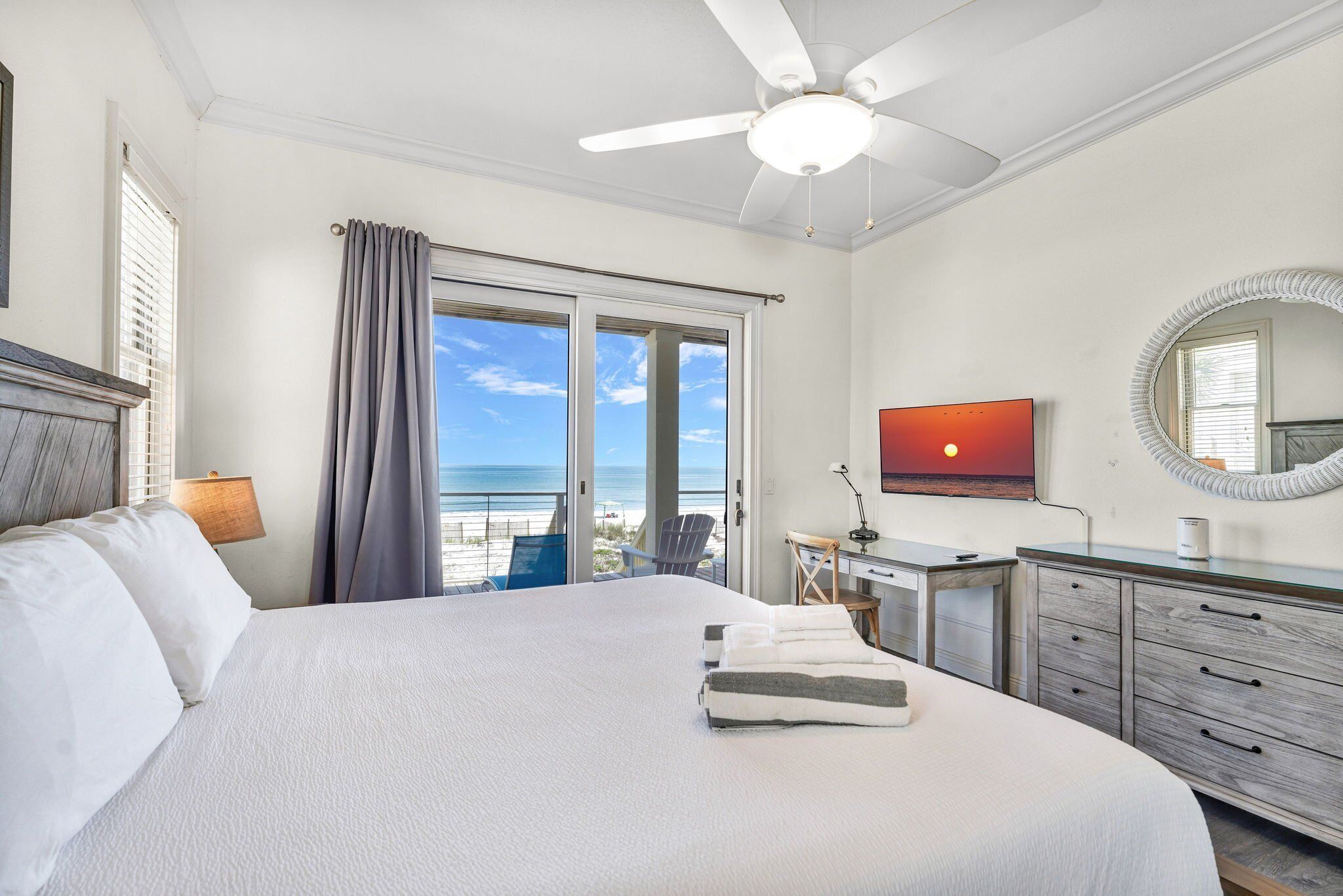 A hotel room with a bed, a desk, a television, and a view of the beach.
