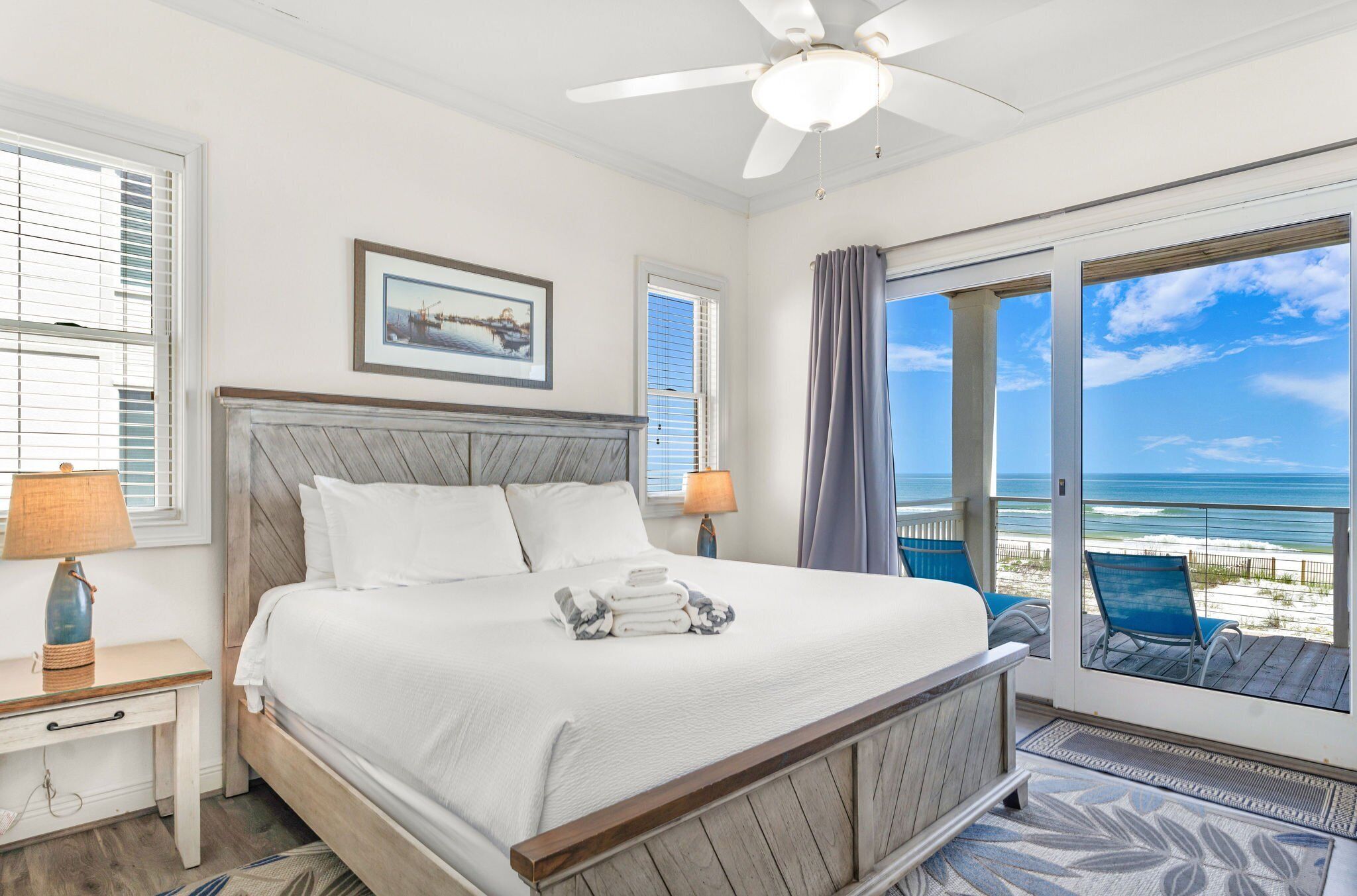 A bedroom with a large bed, a ceiling fan, a window with blinds, a framed picture on the wall, and a view of the beach through a glass door.
