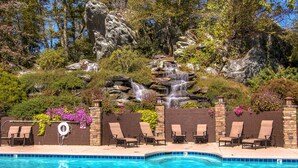 A pool with a waterfall feature, surrounded by lounge chairs and a stone wall with plants. - Escape the Heat in the High Country - 3 King Bedrooms & Amazing Views! (Banner Elk)