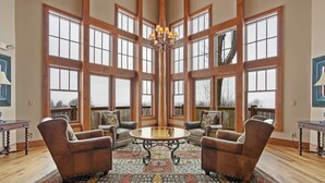 A spacious room with large windows, a chandelier, and leather armchairs around a central table. - Escape the Heat in the High Country - 3 King Bedrooms & Amazing Views! (Banner Elk)