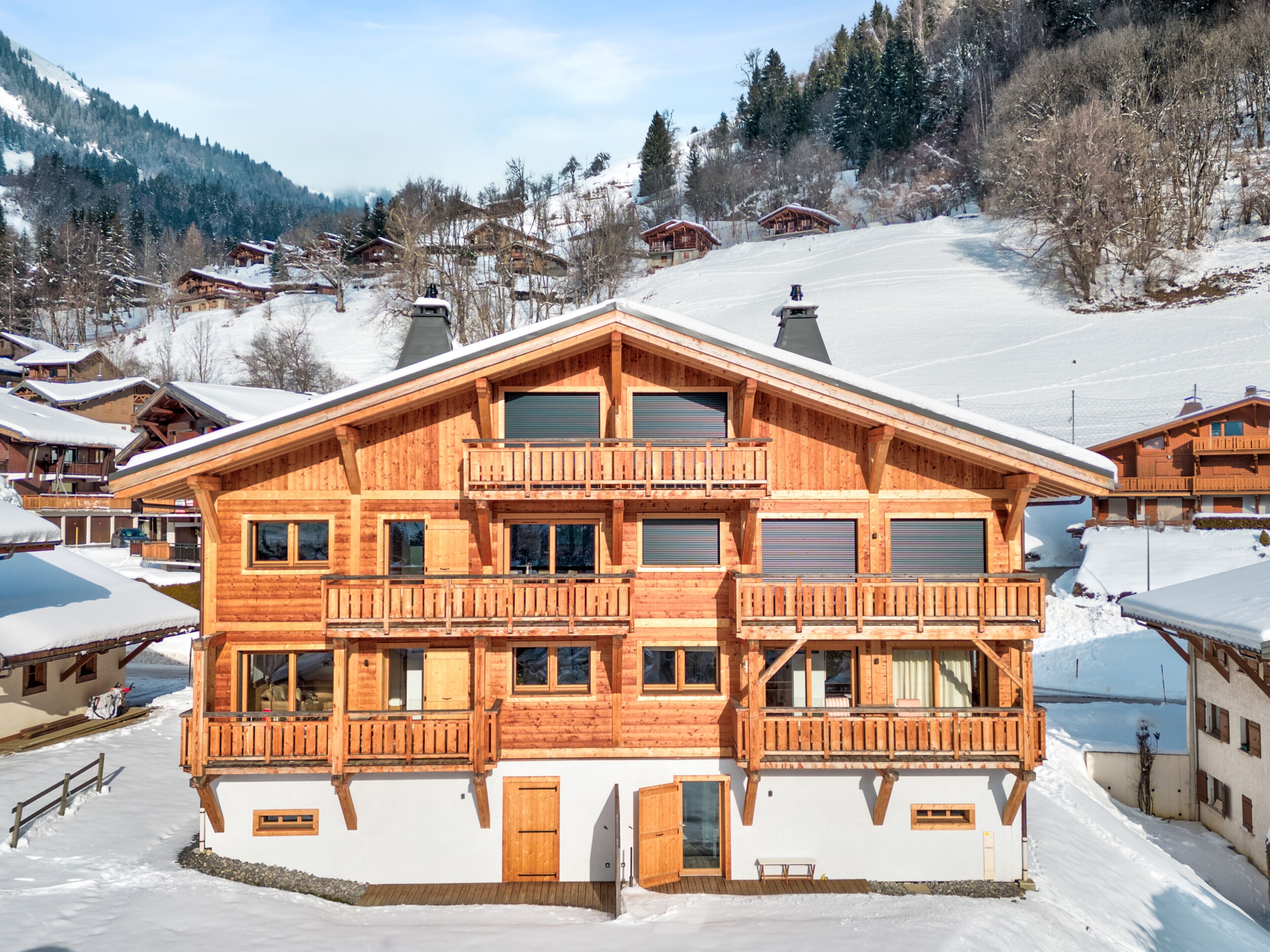 A large wooden chalet with multiple balconies and large windows, surrounded by snow-covered mountains and other chalet-style buildings.
