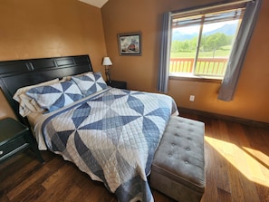 A bedroom with a bed, a nightstand, a window with a view, and a painting on the wall.