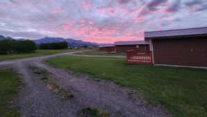 A rural setting with a gravel path leading to wooden cabins, surrounded by greenery and mountains in the background. - Circle R Cabin 1, Private Ranch, Mountain Views (East Glacier Park Village)
