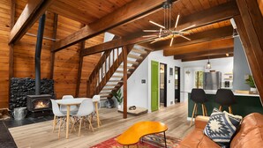 Un loft spacieux avec de hauts plafonds, des poutres apparentes, une cheminée en pierre, un coin repas avec des chaises blanches, une cuisine dotée d’un réfrigérateur moderne et un escalier menant à un niveau supérieur.