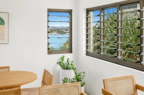 A room with a round wooden table, two wicker chairs, a potted plant, and a window with a view of a body of water and greenery. - Beachfront living & ocean views (Buddina)