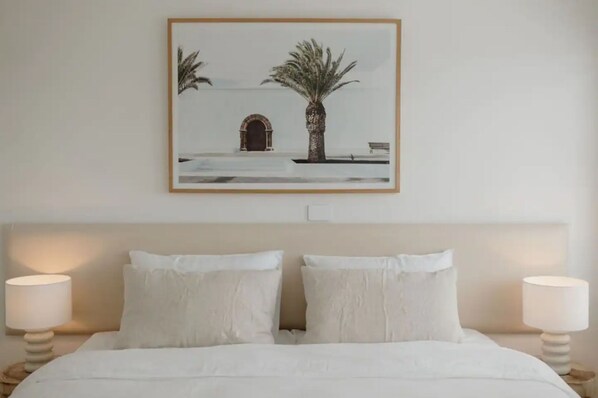 A bedroom with a bed, two bedside lamps, and a framed picture of a palm tree and archway on the wall. - Beachfront living & ocean views (Buddina)
