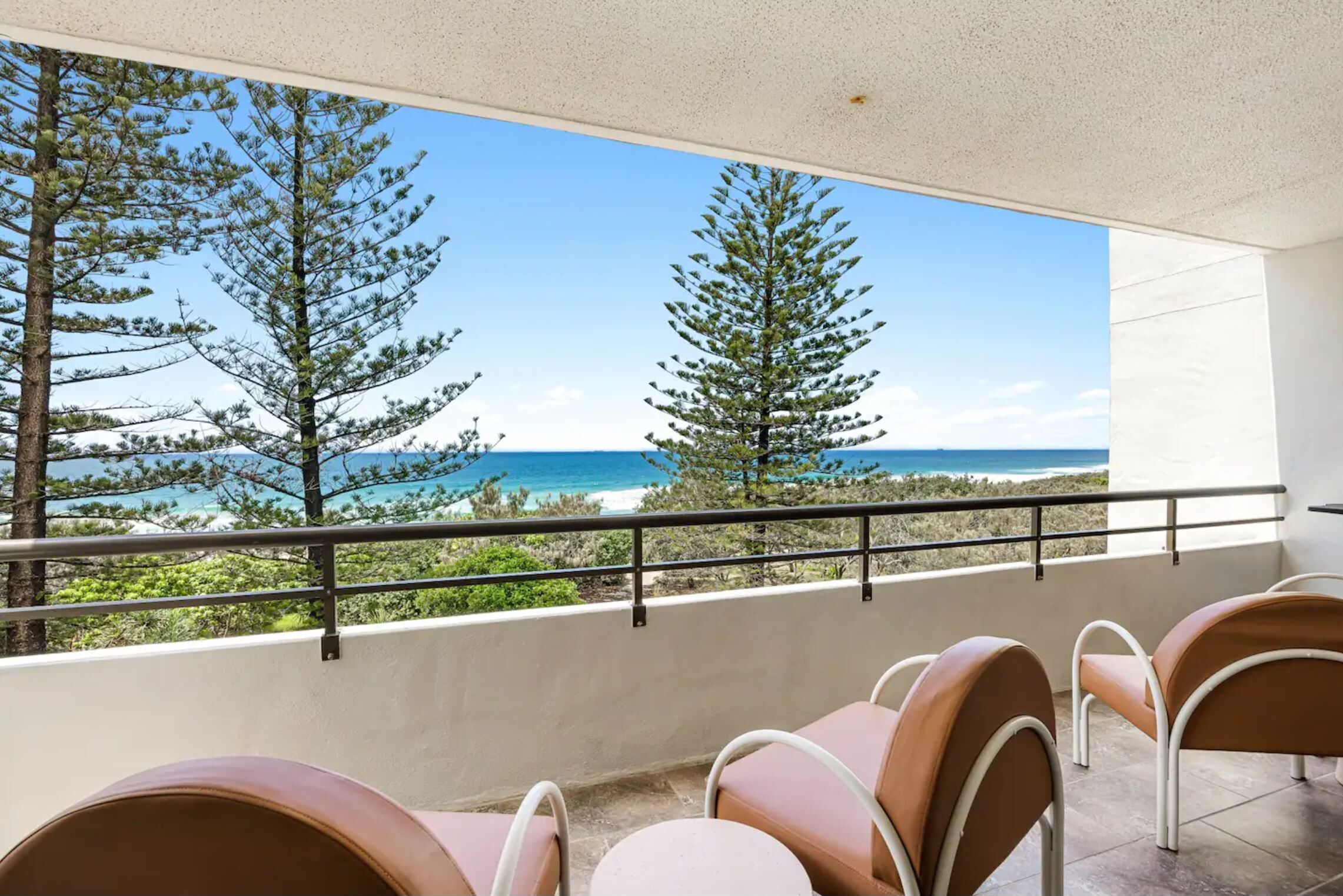A balcony with a view of the ocean and trees, featuring four chairs and a table.