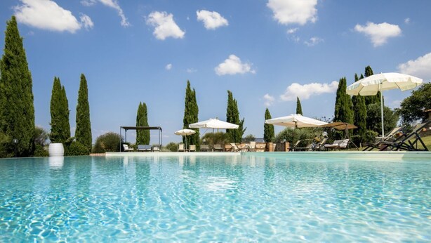 Una piscina cristallina con lettini e ombrelloni, circondata da alberi alti.