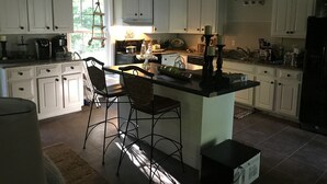 Una cocina con armarios blancos, una isla central con taburetes, una cafetera y un ventanal con luz natural.